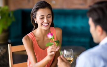 A woman smiles while receiving a pink rose from a person sitting across from her. She is holding a glass of white wine. They are seated at a table in a warmly lit setting with a green velvet sofa in the background.