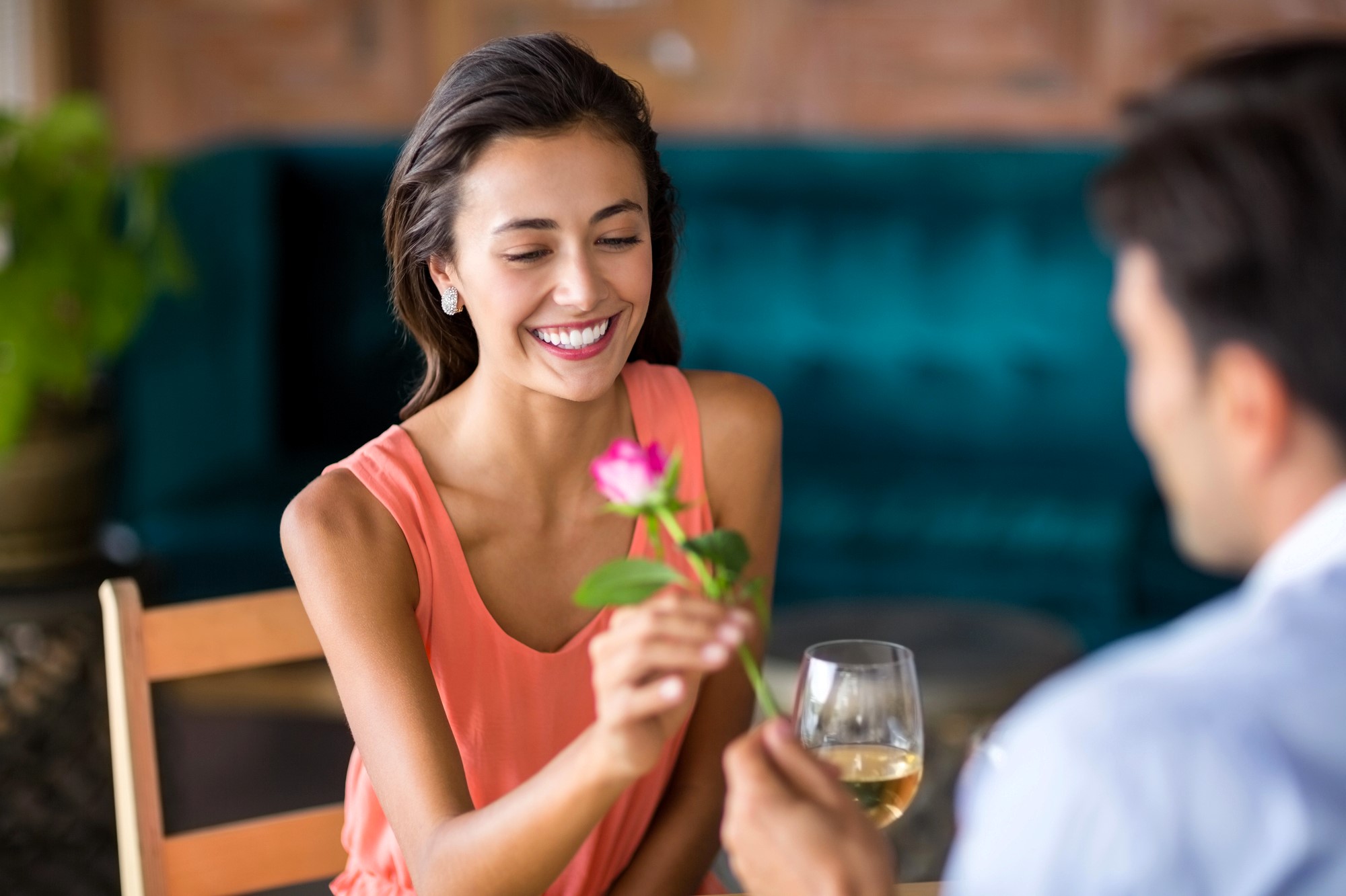 A woman smiles while receiving a pink rose from a person sitting across from her. She is holding a glass of white wine. They are seated at a table in a warmly lit setting with a green velvet sofa in the background.