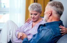 An older couple sits close together on a couch. The woman, with short gray hair, looks down thoughtfully, wearing a striped shirt and white pants. The man, also with gray hair, gazes at her, wearing a denim shirt.