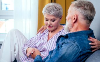 An older couple sits close together on a couch. The woman, with short gray hair, looks down thoughtfully, wearing a striped shirt and white pants. The man, also with gray hair, gazes at her, wearing a denim shirt.