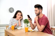 A woman sits at a table, appearing bored while resting her head on her hand, looking at a man beside her. The man is smiling at his phone. They each have a glass of orange juice and a bowl in front of them.