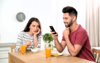 A woman sits at a table, appearing bored while resting her head on her hand, looking at a man beside her. The man is smiling at his phone. They each have a glass of orange juice and a bowl in front of them.