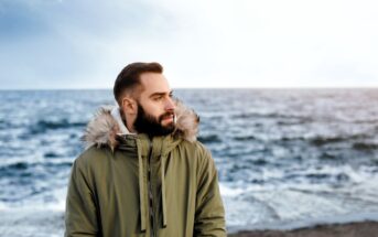 A bearded man in a green parka with a fur-lined hood stands on a beach. He looks to the side, with the ocean in the background under a cloudy sky. The wind appears to be blowing slightly.