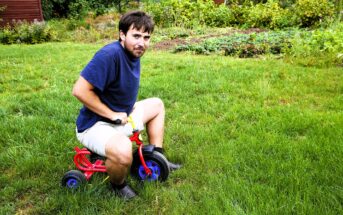 A man with a beard is sitting on a small red tricycle on a grassy lawn. He is wearing a navy blue shirt and beige shorts. The background features green plants and trees.