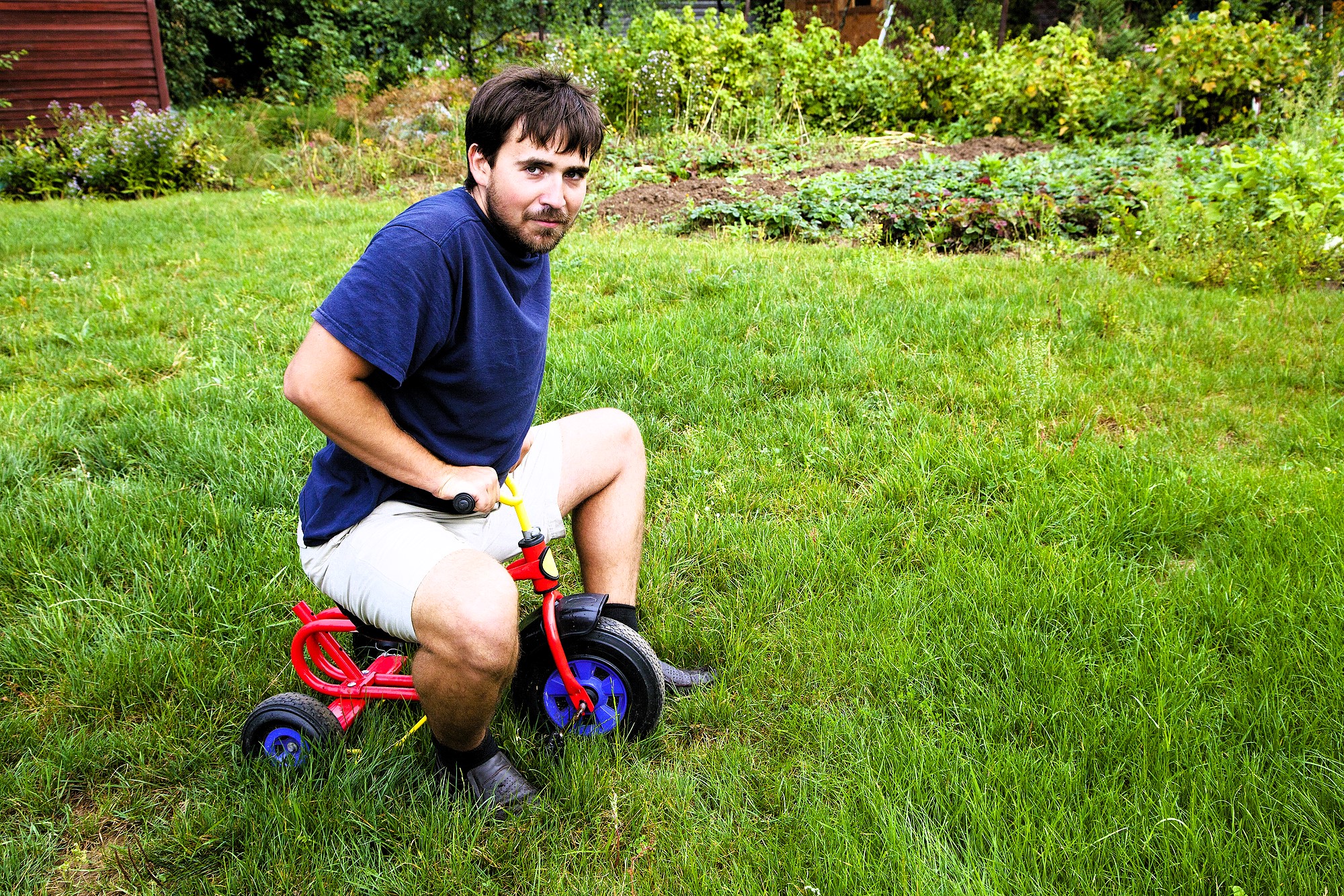 A man with a beard is sitting on a small red tricycle on a grassy lawn. He is wearing a navy blue shirt and beige shorts. The background features green plants and trees.