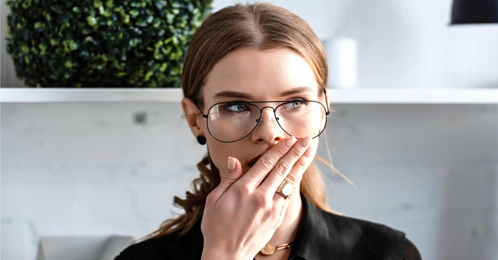 A person with long brown hair wears large round glasses and black clothing, positioned in front of a white background with green foliage. They have their right hand covering their mouth, appearing surprised or concerned.