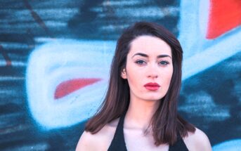 A woman with long brown hair stands in front of a colorful graffiti-covered wall. She wears a sleeveless black top and looks directly at the camera with a neutral expression. The graffiti features red, white, and blue shapes.