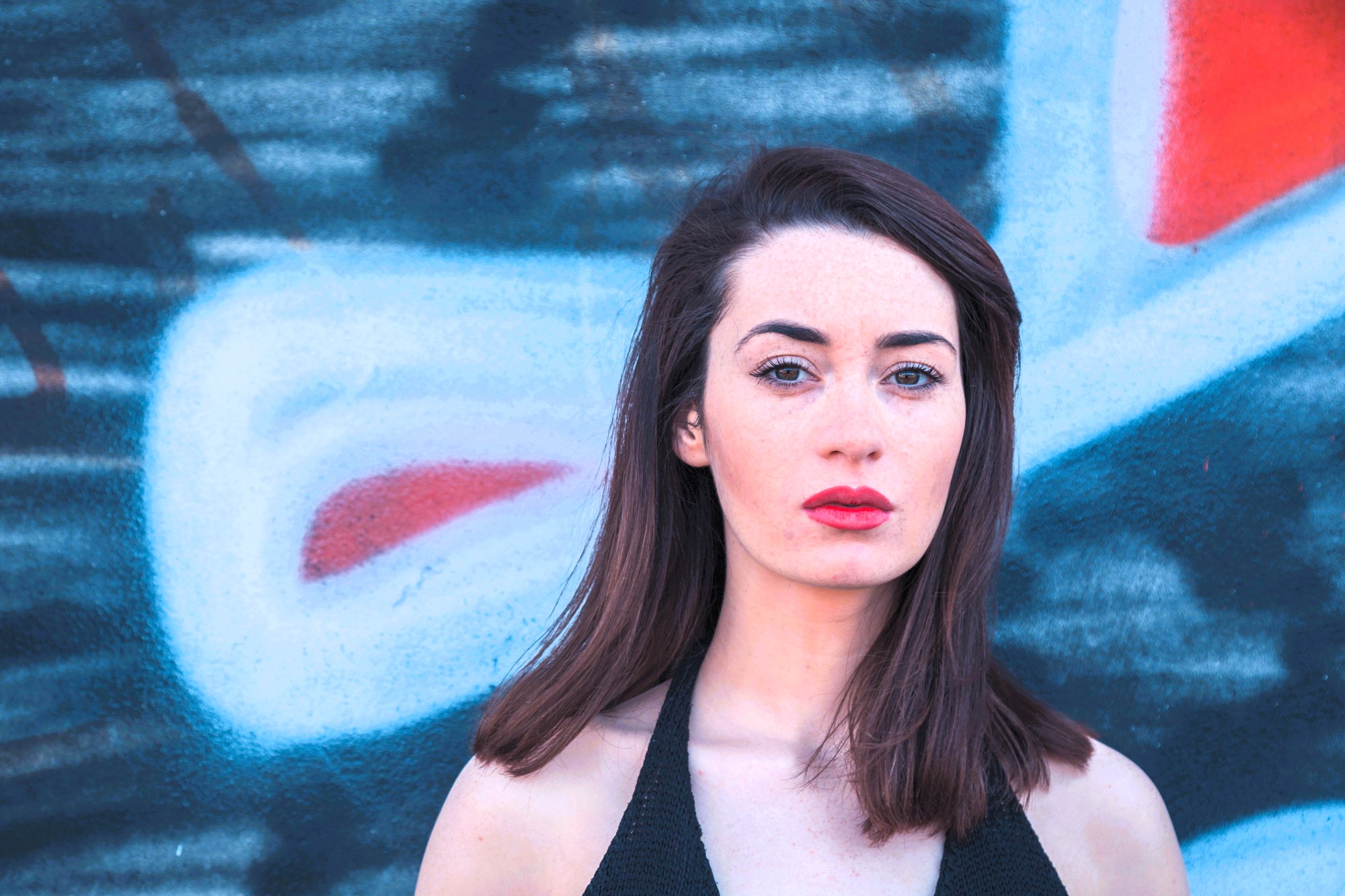 A woman with long brown hair stands in front of a colorful graffiti-covered wall. She wears a sleeveless black top and looks directly at the camera with a neutral expression. The graffiti features red, white, and blue shapes.