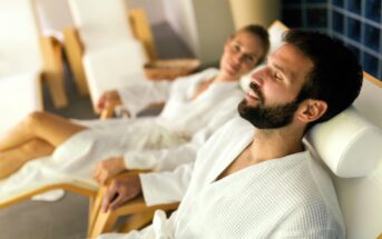 A man and woman are relaxing on reclined chairs in a spa. Both are wearing white bathrobes and appear content. The man has his eyes closed, while the woman looks towards the camera. The setting is calm and peaceful.