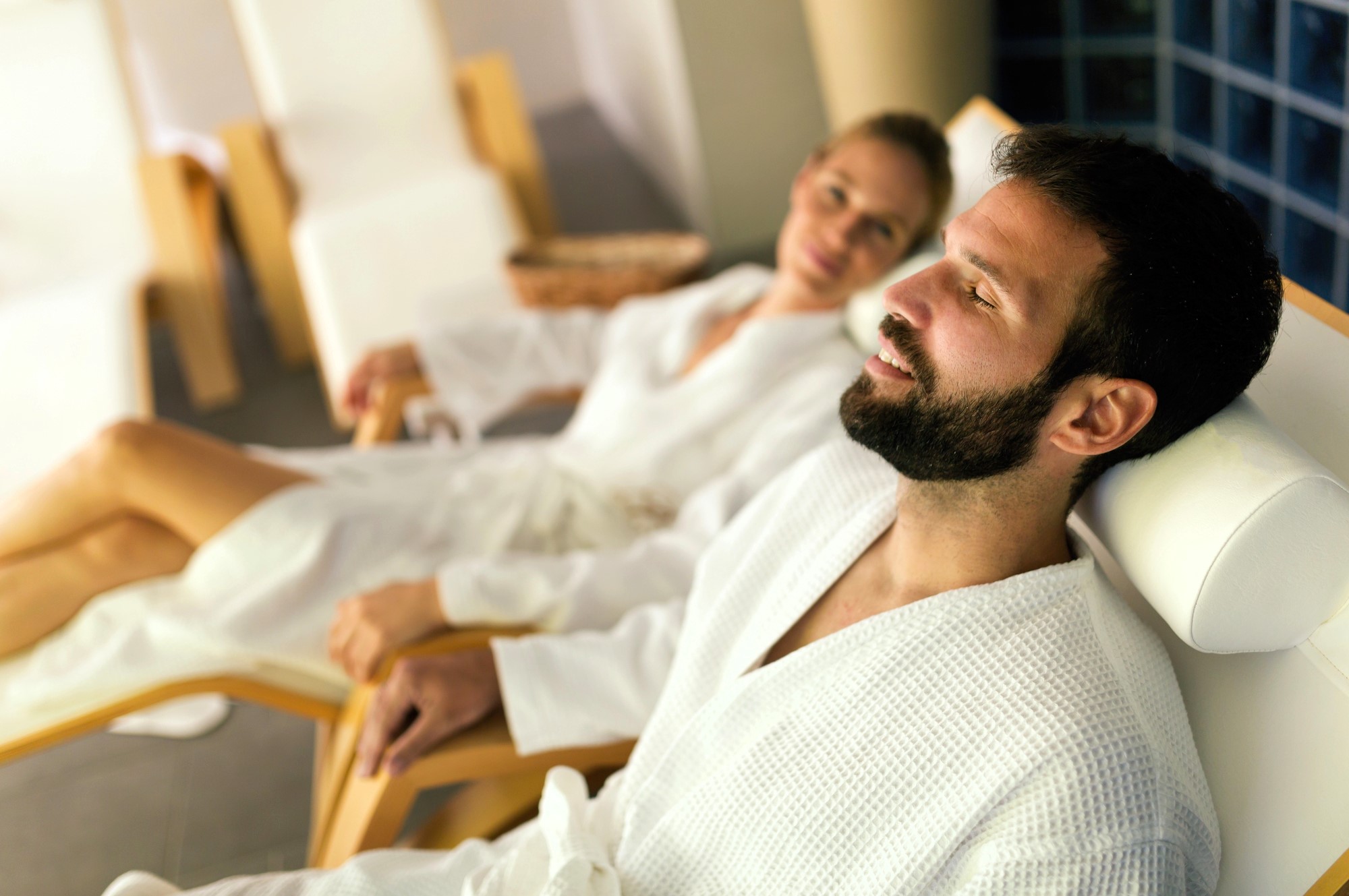 A man and woman are relaxing on reclined chairs in a spa. Both are wearing white bathrobes and appear content. The man has his eyes closed, while the woman looks towards the camera. The setting is calm and peaceful.