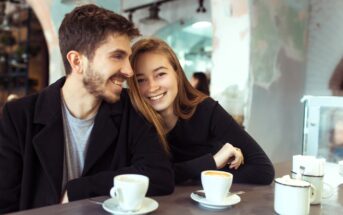 A smiling couple sits at a café table with cups of coffee. The man has short dark hair and a beard, wearing a black coat. The woman has long light brown hair and leans on the table, wearing a black top. The café interior is softly lit.