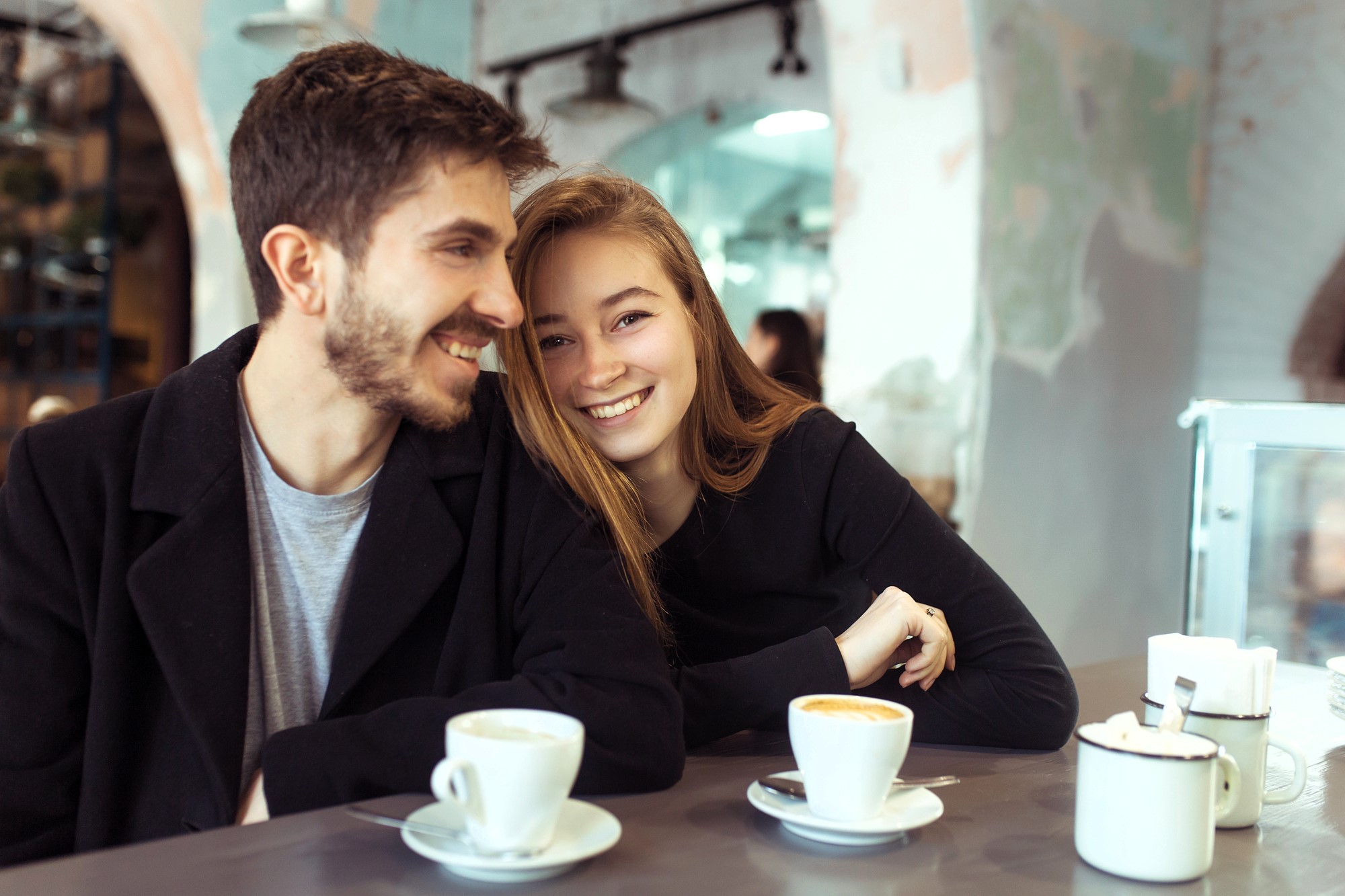 A smiling couple sits at a café table with cups of coffee. The man has short dark hair and a beard, wearing a black coat. The woman has long light brown hair and leans on the table, wearing a black top. The café interior is softly lit.