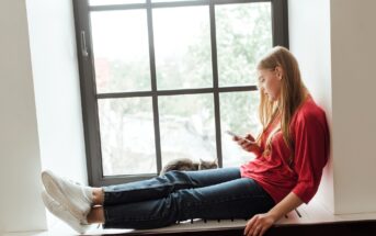 A young woman in a red shirt and jeans sits on a windowsill, looking at her phone. A gray cat rests beside her. The large window behind them shows a blurred view of greenery outside.