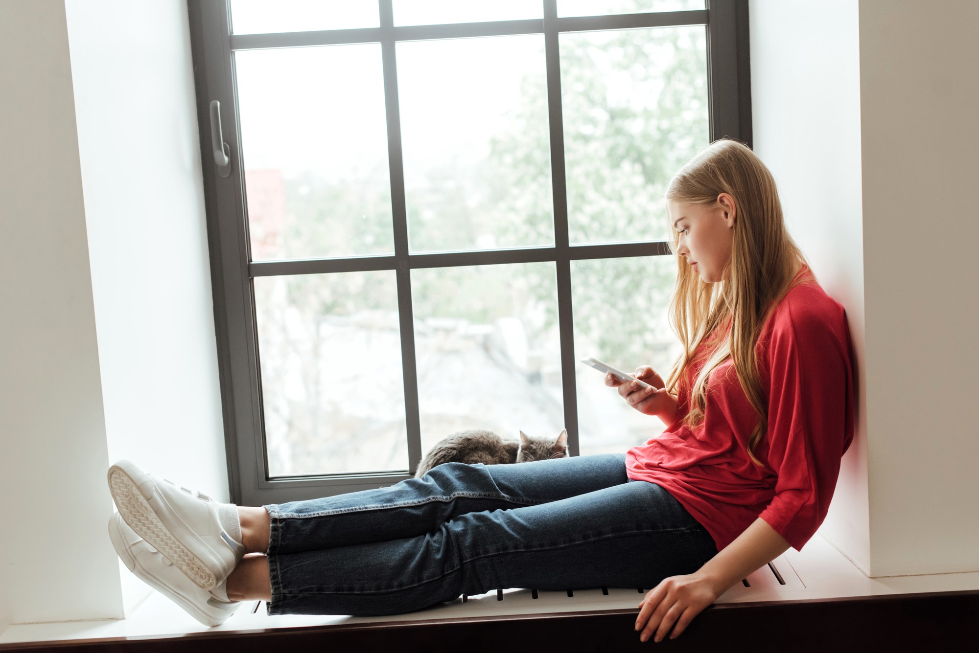 A young woman in a red shirt and jeans sits on a windowsill, looking at her phone. A gray cat rests beside her. The large window behind them shows a blurred view of greenery outside.