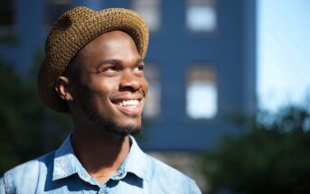 A person wearing a straw hat and a denim shirt smiles while looking upwards. The background is a blurred urban setting with sunlight casting a warm glow.