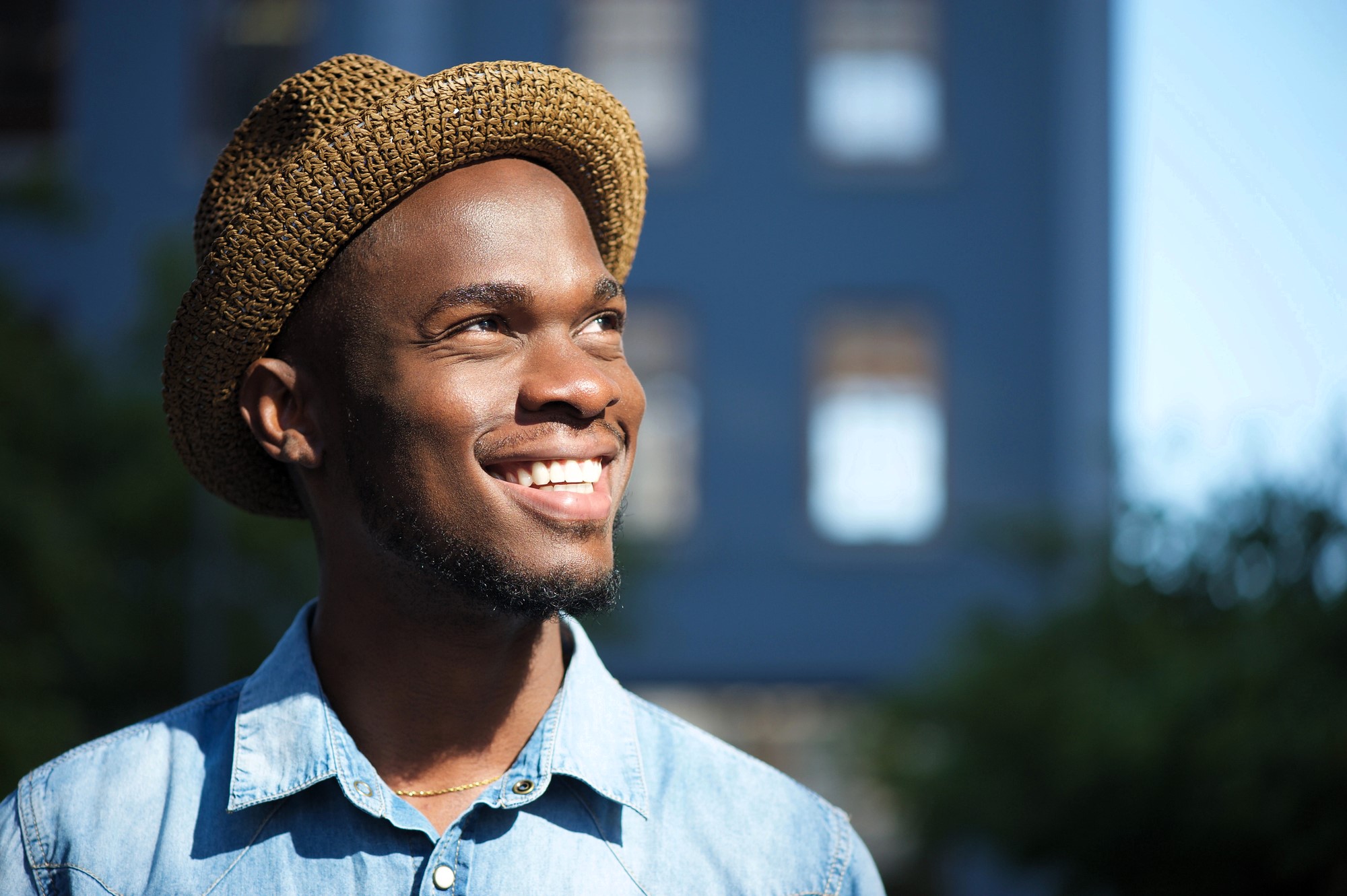 A person wearing a straw hat and a denim shirt smiles while looking upwards. The background is a blurred urban setting with sunlight casting a warm glow.