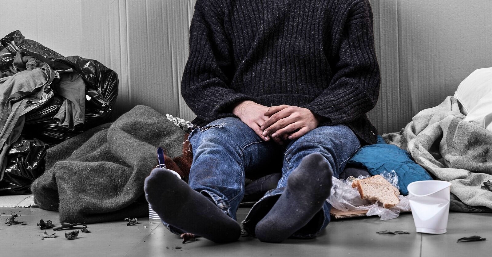 homeless person from the neck down sitting on the floor surrounded by his possessions