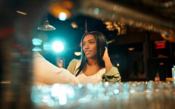 Woman with long hair sits at a bar, smiling while looking to the side. The background is softly blurred with colorful lights, creating a warm, inviting atmosphere. Glasses are visible in the foreground.