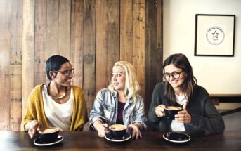 Three people sit at a wooden table in a cafe, each holding a cup of coffee. They are smiling and engaged in conversation. A framed picture hangs on the wooden wall behind them.