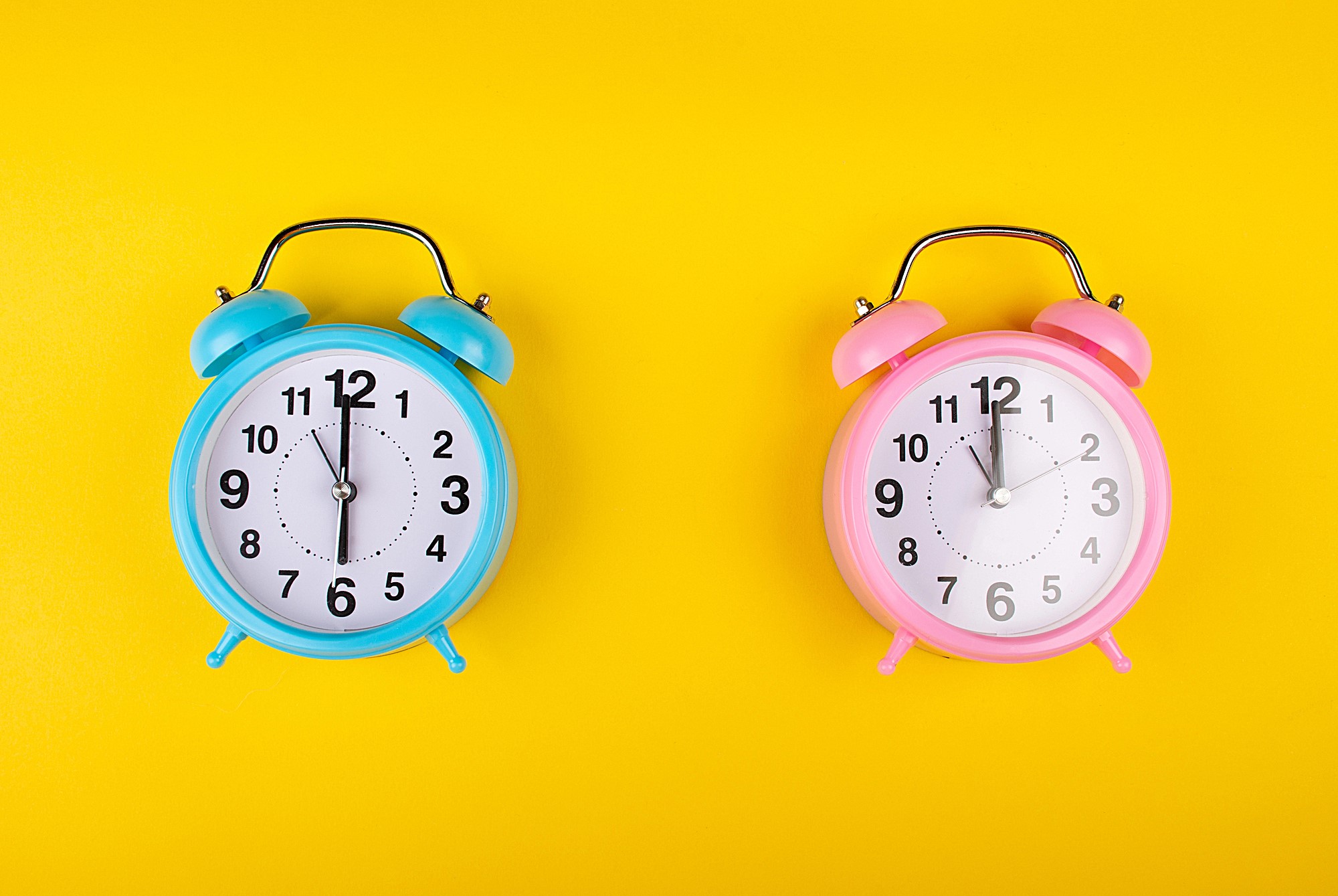 Two alarm clocks on a yellow background: one blue on the left showing 10:09 and one pink on the right showing 3:54. Both clocks have classic twin bell designs with metal handles.