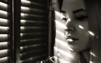 A woman gazes downward, with sunlight streaming through window blinds casting stripes of light and shadow across her face. The scene is in black and white, creating a contemplative and moody atmosphere.