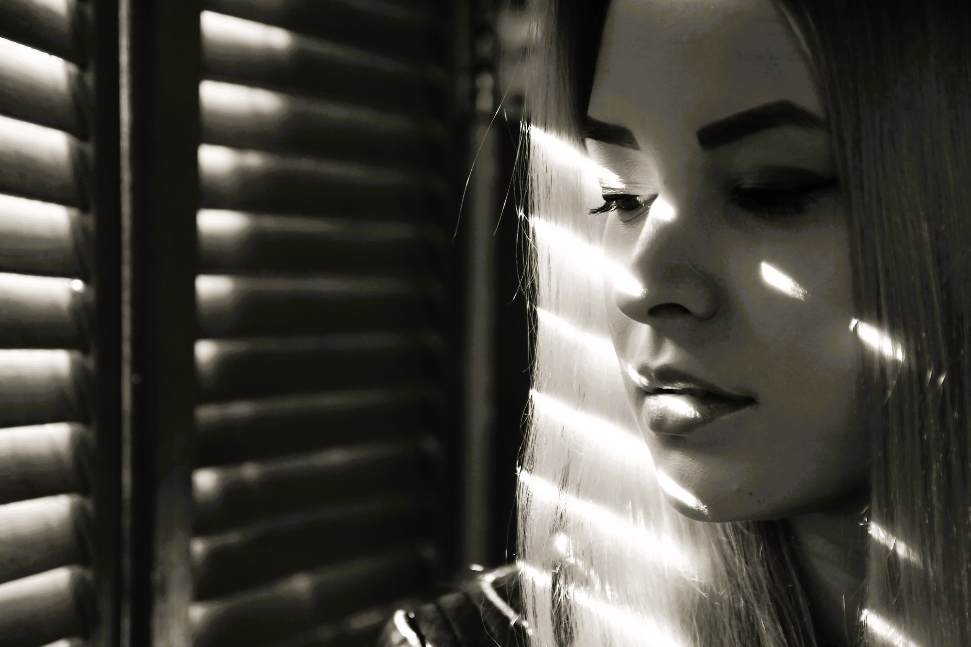 A woman gazes downward, with sunlight streaming through window blinds casting stripes of light and shadow across her face. The scene is in black and white, creating a contemplative and moody atmosphere.