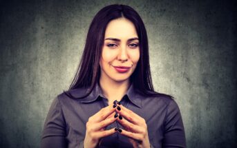 Woman with long dark hair in a gray blouse, smirking and clasping hands together with fingertips touching, stands against a textured gray background.