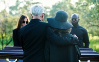 A group of people dressed in black stand by a coffin outdoors during a funeral. The central figure, an older man, has his arm around a woman wearing a large hat. Two other individuals are present, with blurred greenery in the background.