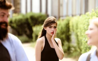 A woman in a black top covers her mouth with her hand, looking startled or concerned. She stands outdoors on a sunlit pathway with blurred greenery in the background. Two other people, out of focus, are in the foreground.