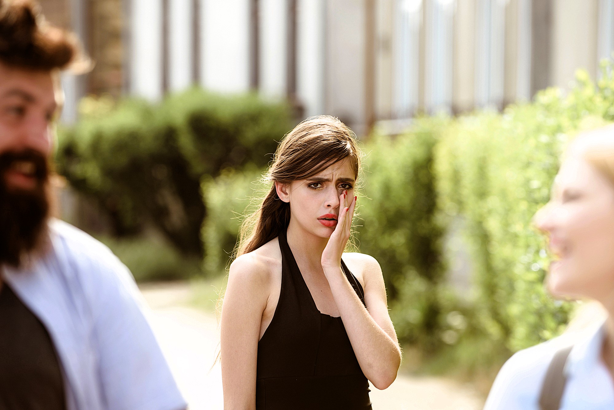 A woman in a black top covers her mouth with her hand, looking startled or concerned. She stands outdoors on a sunlit pathway with blurred greenery in the background. Two other people, out of focus, are in the foreground.