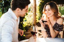 A man and woman are sitting at an outdoor table, enjoying glasses of red wine. They are smiling at each other, surrounded by lush greenery. The woman is wearing an off-shoulder floral dress, and the man is in a white shirt.