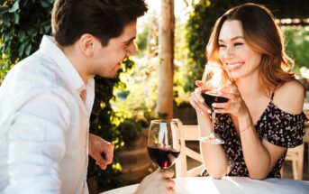 A man and woman are sitting at an outdoor table, enjoying glasses of red wine. They are smiling at each other, surrounded by lush greenery. The woman is wearing an off-shoulder floral dress, and the man is in a white shirt.