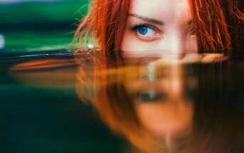 A close-up of a person with red hair partially submerged in water. They have one visible blue eye peeking above the water surface, with their reflection mirrored on the water below. The background is a blurred mix of green and blue hues.