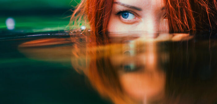 A close-up of a person with red hair partially submerged in water. They have one visible blue eye peeking above the water surface, with their reflection mirrored on the water below. The background is a blurred mix of green and blue hues.