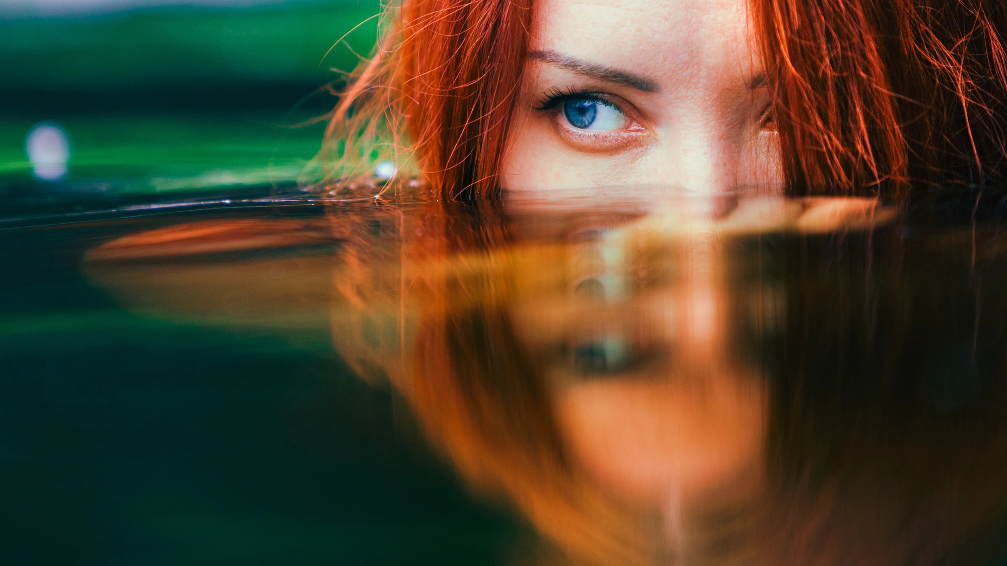 A close-up of a person with red hair partially submerged in water. They have one visible blue eye peeking above the water surface, with their reflection mirrored on the water below. The background is a blurred mix of green and blue hues.