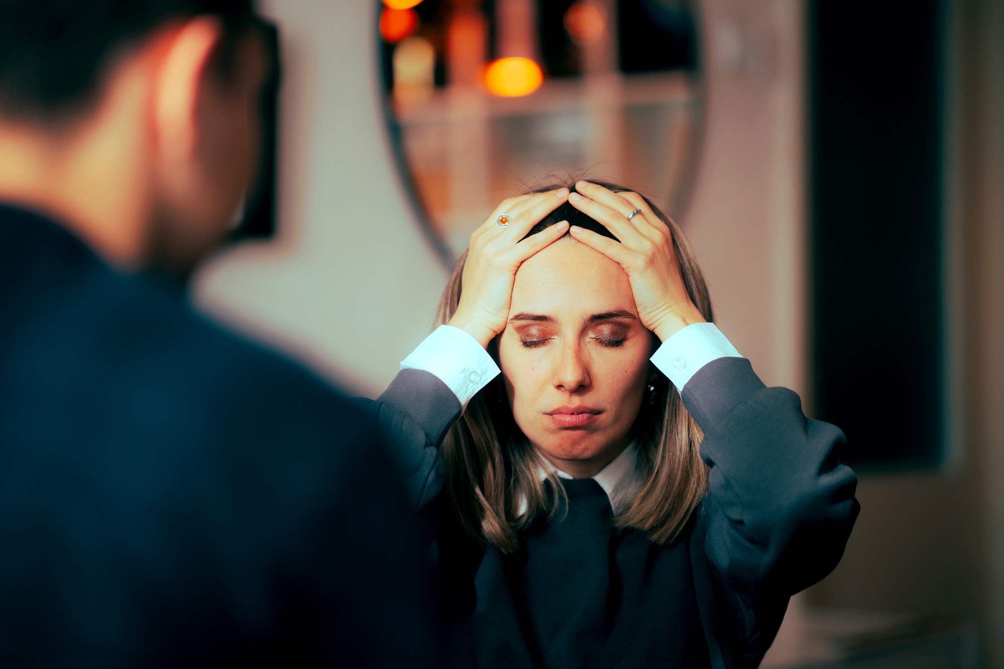 A woman with blonde hair sits facing a man, holding her head in her hands with a distressed expression. She is indoors, with a blurred background featuring warm lighting and circular decorations on the wall.