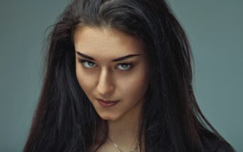 A woman with long, dark hair and intense blue eyes stares directly into the camera. She has a slight smile on her face and wears a silver necklace. The background is a muted gray-blue, which contrasts with her dark hair.