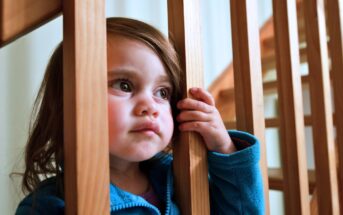 A young child with long brown hair and wearing a blue jacket looks thoughtfully through wooden stair railings, holding one rail with her hand. The background is softly lit, highlighting the wooden texture of the stairs.