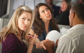 A woman with blonde hair in the foreground is making a skeptical face and pointing at a man with short hair. A woman with brown hair in the background has a confused expression. Three other people are partially visible, engaged in conversation.