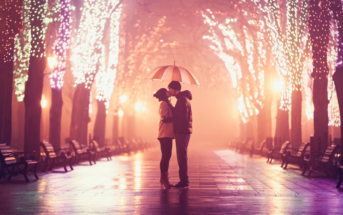 A couple stands under an umbrella, sharing an intimate moment in the center of a path lined with trees adorned with sparkling lights. The glow creates a magical, romantic atmosphere against the misty background. Empty benches line the sides of the path.