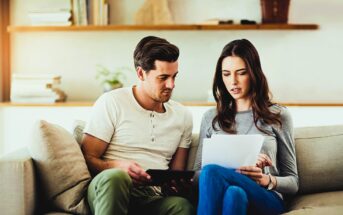 A man and a woman sit on a beige couch in a living room. The man holds a tablet, while the woman looks at documents. They appear to be discussing something. A wooden shelf with books and decor is visible in the background.