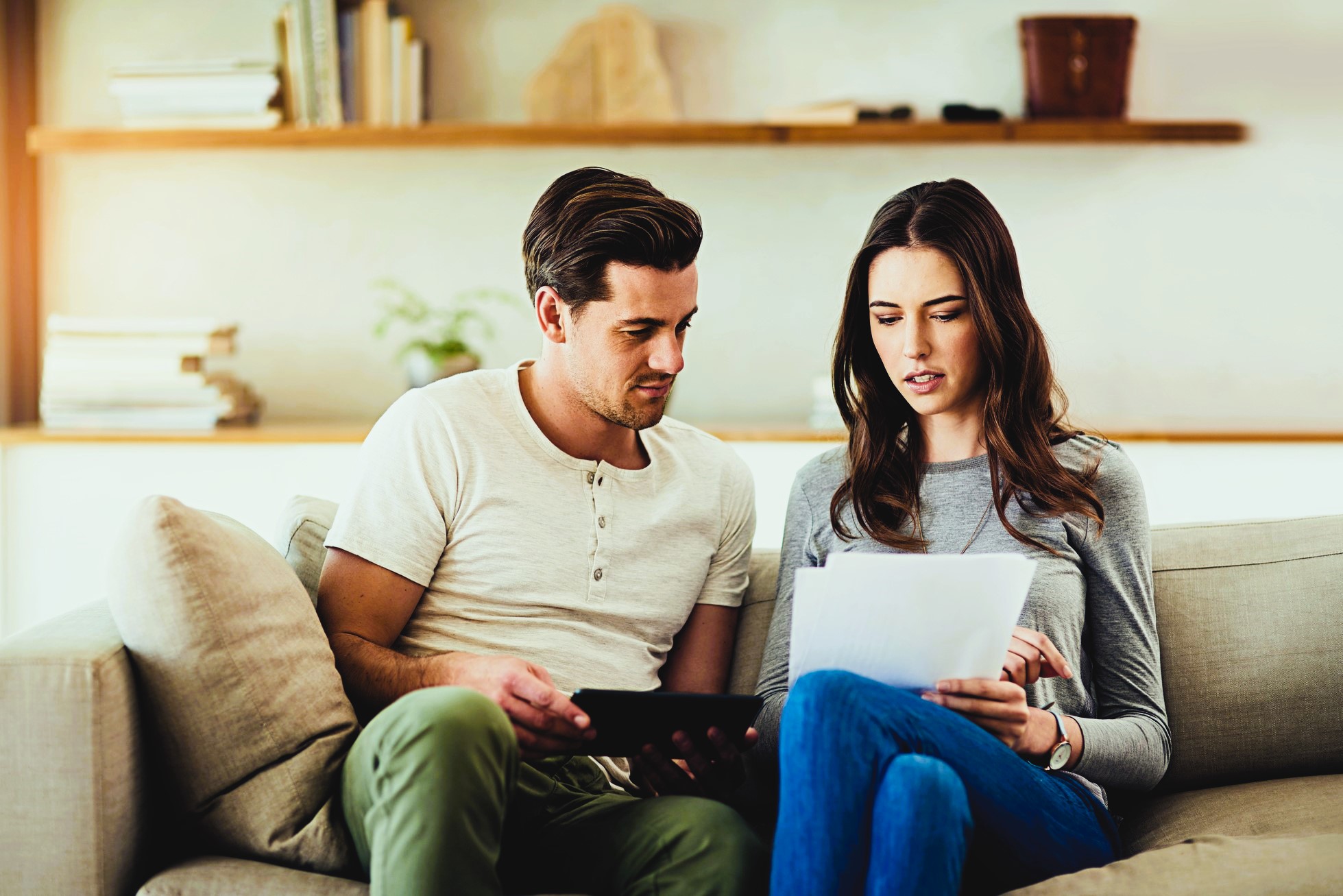 A man and a woman sit on a beige couch in a living room. The man holds a tablet, while the woman looks at documents. They appear to be discussing something. A wooden shelf with books and decor is visible in the background.