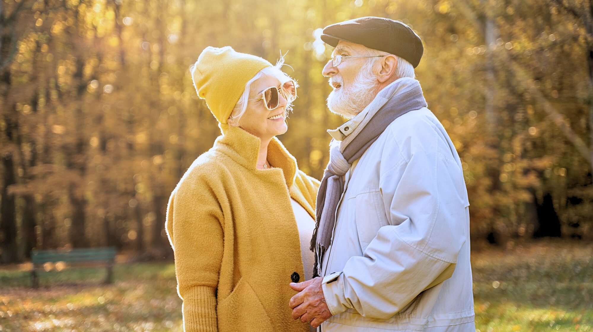 An elderly couple is standing outside in a sunlit autumn park. The woman is wearing a yellow coat, matching beanie, and sunglasses, while the man is dressed in a light-colored jacket, scarf, and beret. They are smiling and gazing at each other affectionately.