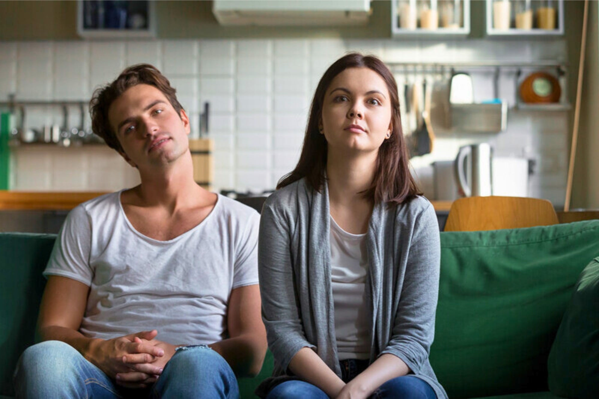 A man and a woman sit on a green couch in a room with a kitchen in the background. The man, wearing a white t-shirt, leans back with a relaxed expression. The woman, in a gray cardigan and white shirt, sits upright, looking ahead.