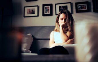 A woman sits on a couch with a pensive expression, resting her chin on her hand. The background features several framed photos on the wall. The lighting is soft, creating a cozy and reflective atmosphere.