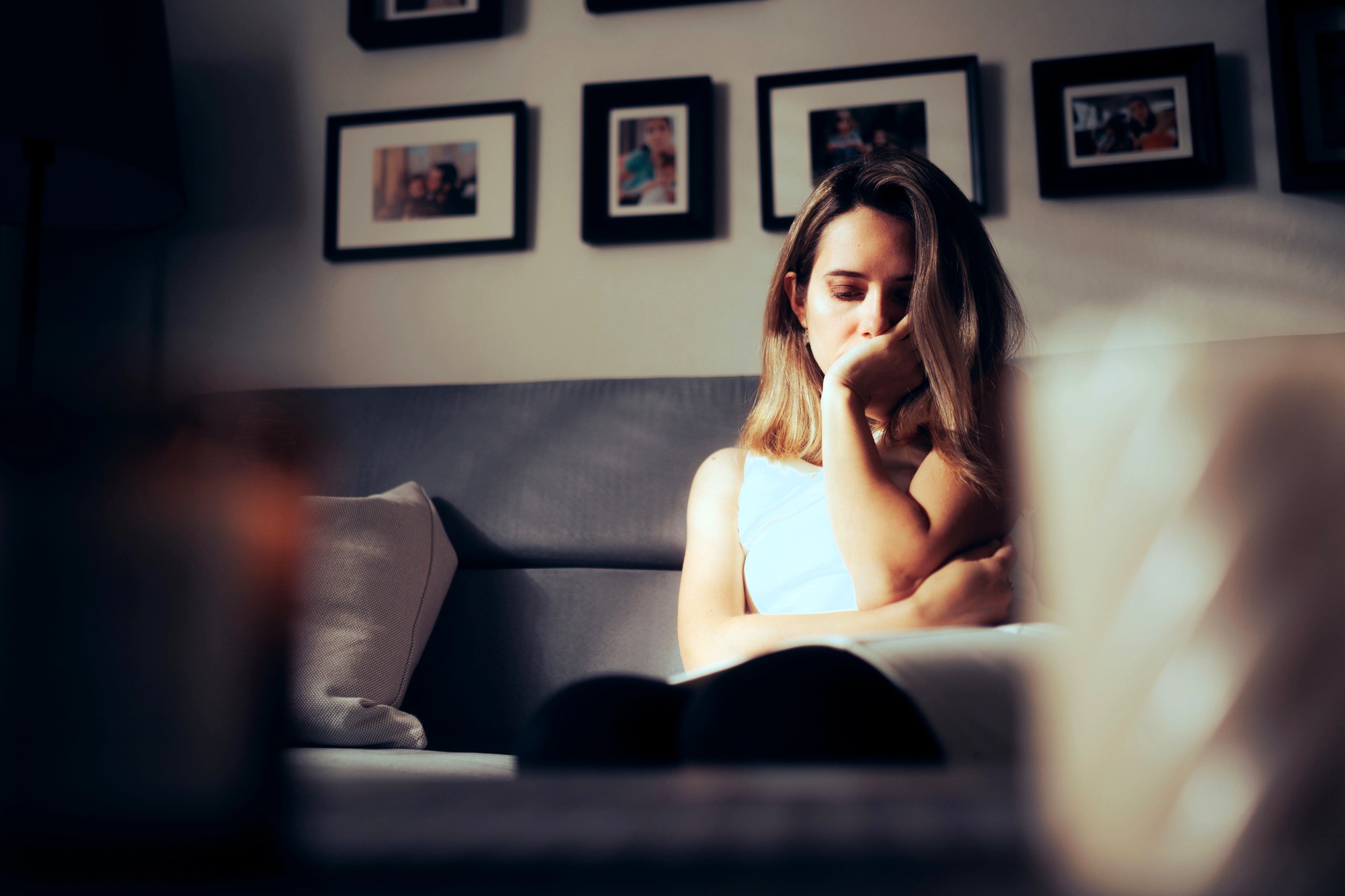 A woman sits on a couch with a pensive expression, resting her chin on her hand. The background features several framed photos on the wall. The lighting is soft, creating a cozy and reflective atmosphere.