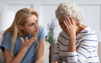 A young woman with blonde hair is sitting next to an older woman with curly gray hair. The older woman appears distressed, resting her head in her hand. The young woman is gesturing and seems to be speaking, showing concern. They are indoors, seated on a couch.