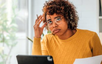 A person with curly hair and glasses, wearing a yellow sweater, sits at a desk. They hold a pen and a sheet of paper, looking confused or frustrated, with a tablet in front of them. Bright, blurred background with natural light.