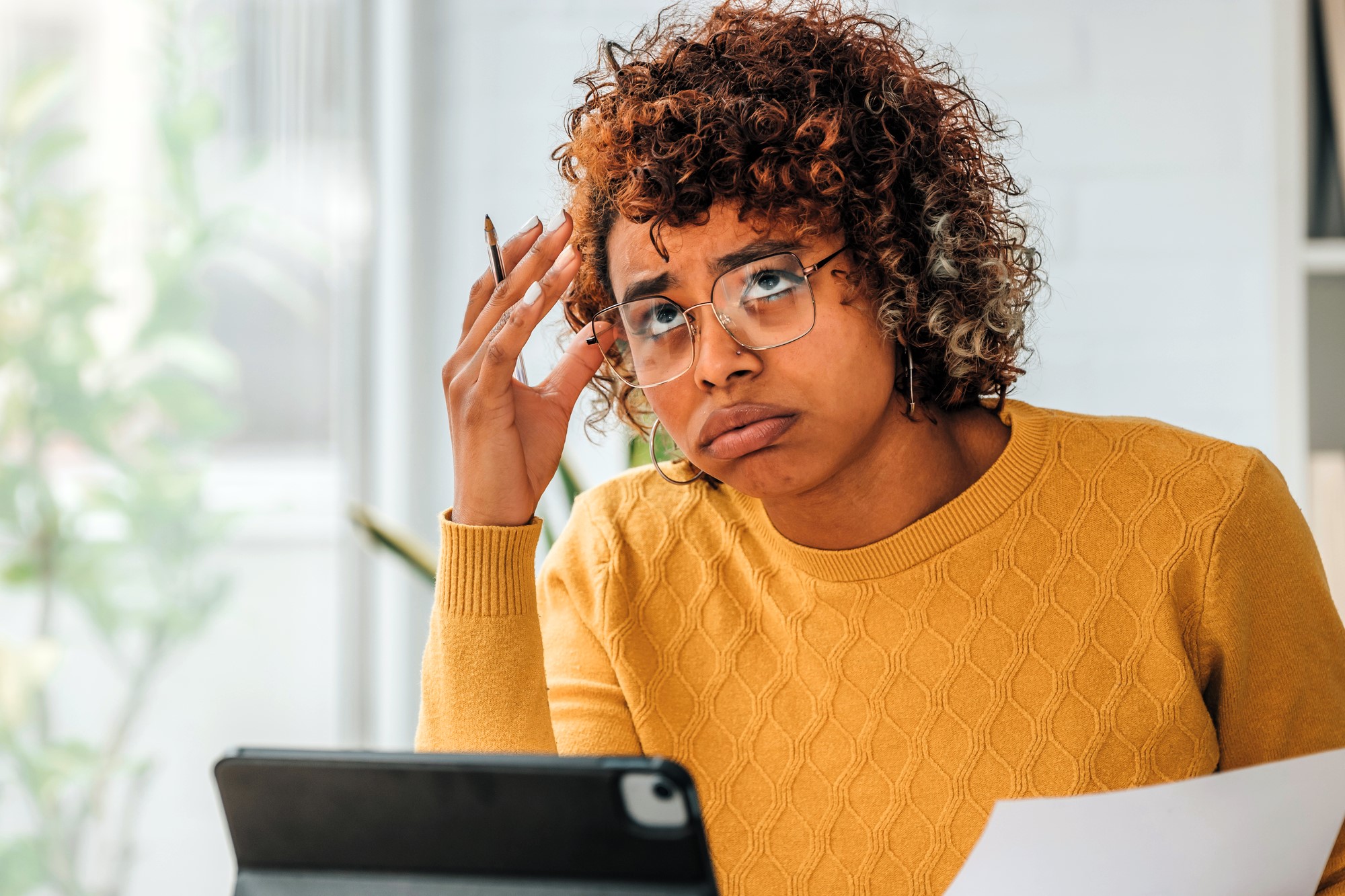 A person with curly hair and glasses, wearing a yellow sweater, sits at a desk. They hold a pen and a sheet of paper, looking confused or frustrated, with a tablet in front of them. Bright, blurred background with natural light.