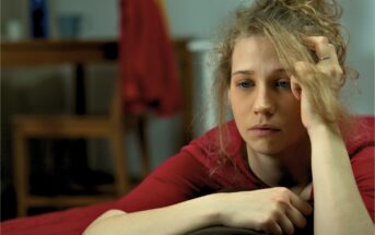 A young woman with blonde curly hair rests her head on her hand, looking contemplative and slightly sad. She wears a red top and is leaning on a surface with a blurred background that includes a chair and a red garment hanging.